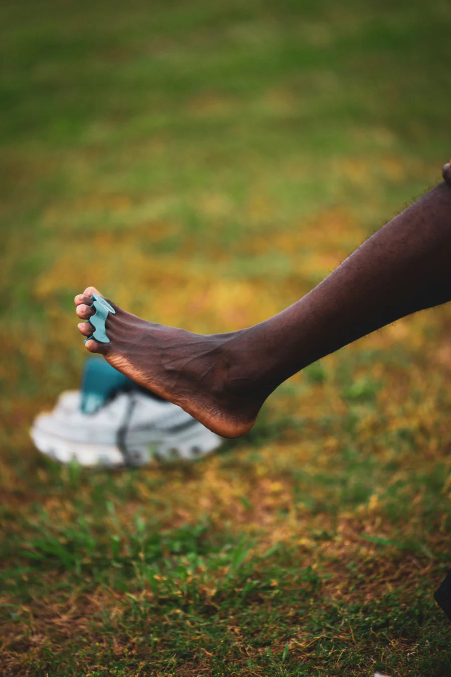 Barfota fot bär en turkos tåseparator från Correct Toes, lyft i luften framför en gräsbeklädd bakgrund. En sportskosula syns i bakgrunden, vilket antyder användning i naturlig miljö för fotträning eller återhämtning.
